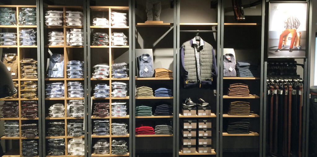 Blick auf eine Modesteppe mit verschiedenen Herrenbekleidungsartikeln. Links sind Regale mit eingepackten, gestapelten Hemden, T-Shirts und Pullovern. In der Mitte hängen Jacken und Mäntel, darunter sind verschiedene gestapelte Pullover in neutralen und dunklen Farben. Rechts sind Kleidung und Schuhe in separaten Fächern sichtbar, darunter auch Gürtel.