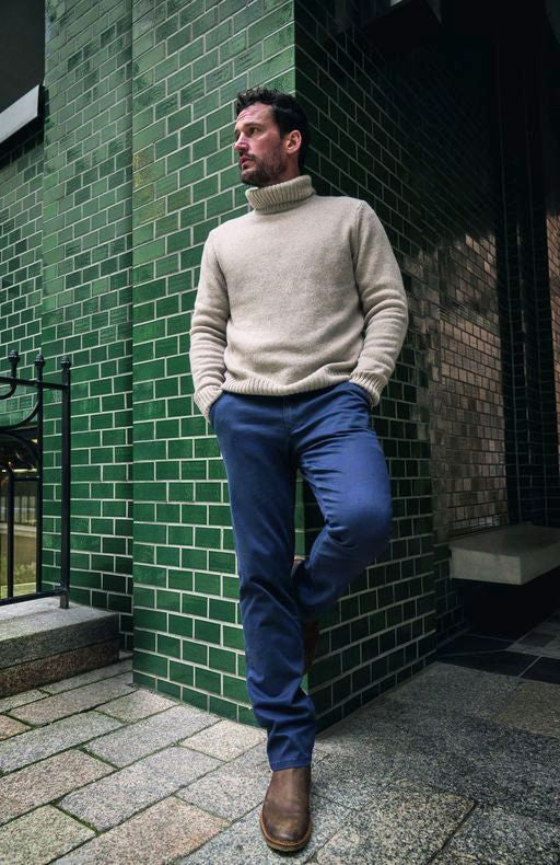 Ein Mann mit dunklem Haart und Bart steht an einer Ecke einer grünen Ziegelwand auf einer Straßenkreuzung. Er trägt einen hellgrauen Rollkragenpullover, blaue Hosen und braune Stiefel. Seine Hände sind in den Taschen, er blickt nach rechts in die Ferne.