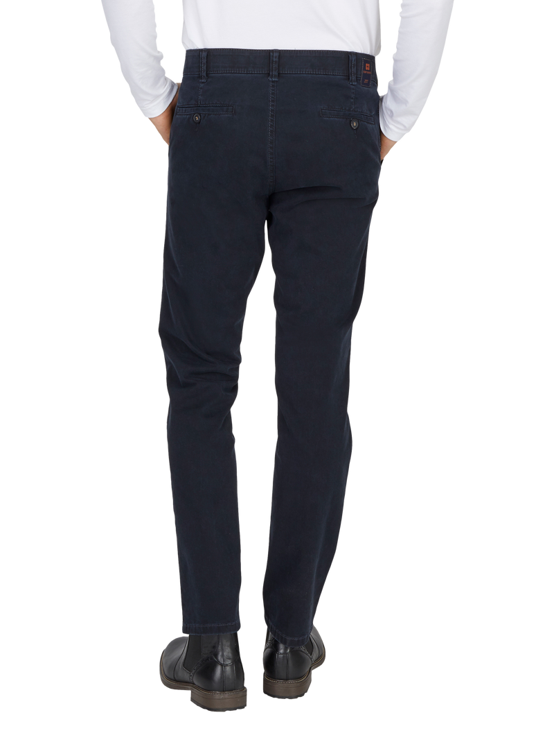 Blick auf einen Mann, der dunkelblaue, schmale Chino-Hose trägt, die hinten sichtbar ist. Er hat die Hände in den Taschen, trägt eine weiße Hemdjacke und schwarze Schuhe.