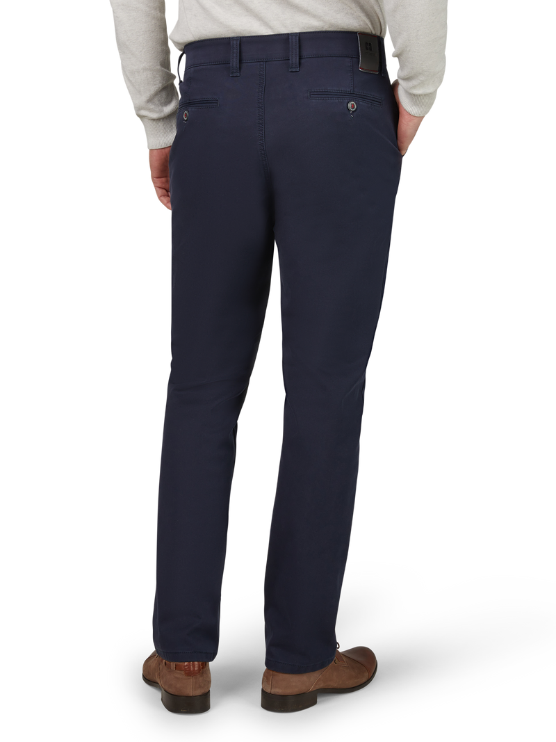 Hochgezogene schwarze Anzughose aus festem Stoff, getragen von einer Person, die eine helle Bluse und braune Schuhe trägt. Die Hose hat Gürtelschlaufen, zwei hintere Einschubtaschen mit Knopfverschluss und eine lederne Markenetikette am Bund. Die Person steht mit den Händen in den Taschen, Blick nach hinten gerichtet.