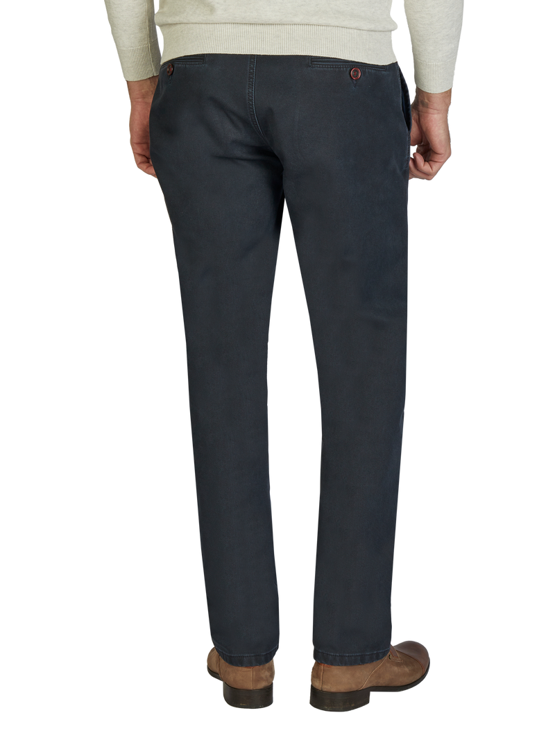 Ein Mann trägt dunkelblaue Baumwollhosen, die locker sitzen und vorne ein Reißverschluss sowie Knöpfe oben haben. Die Hose hat seitliche Taschen und sitzt auf Hüfthöhe. Er trägt braune Schuhe und ein helles Oberteil.