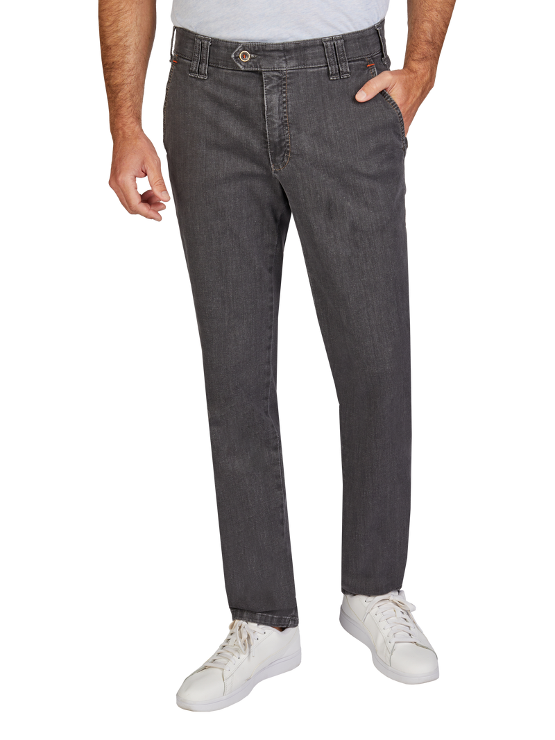 Blick auf eine Person, die dunkle, geradlinige Jeans trägt, mit einer Hand in der Tasche. Die Jeans hat einen klassischen Schnitt, eine Gürtelschlaufe und eine Metallknopf-Verschluss. Die Person trägt weiße Schuhe und ein hellgraues Oberteil, das nur teilweise sichtbar ist.