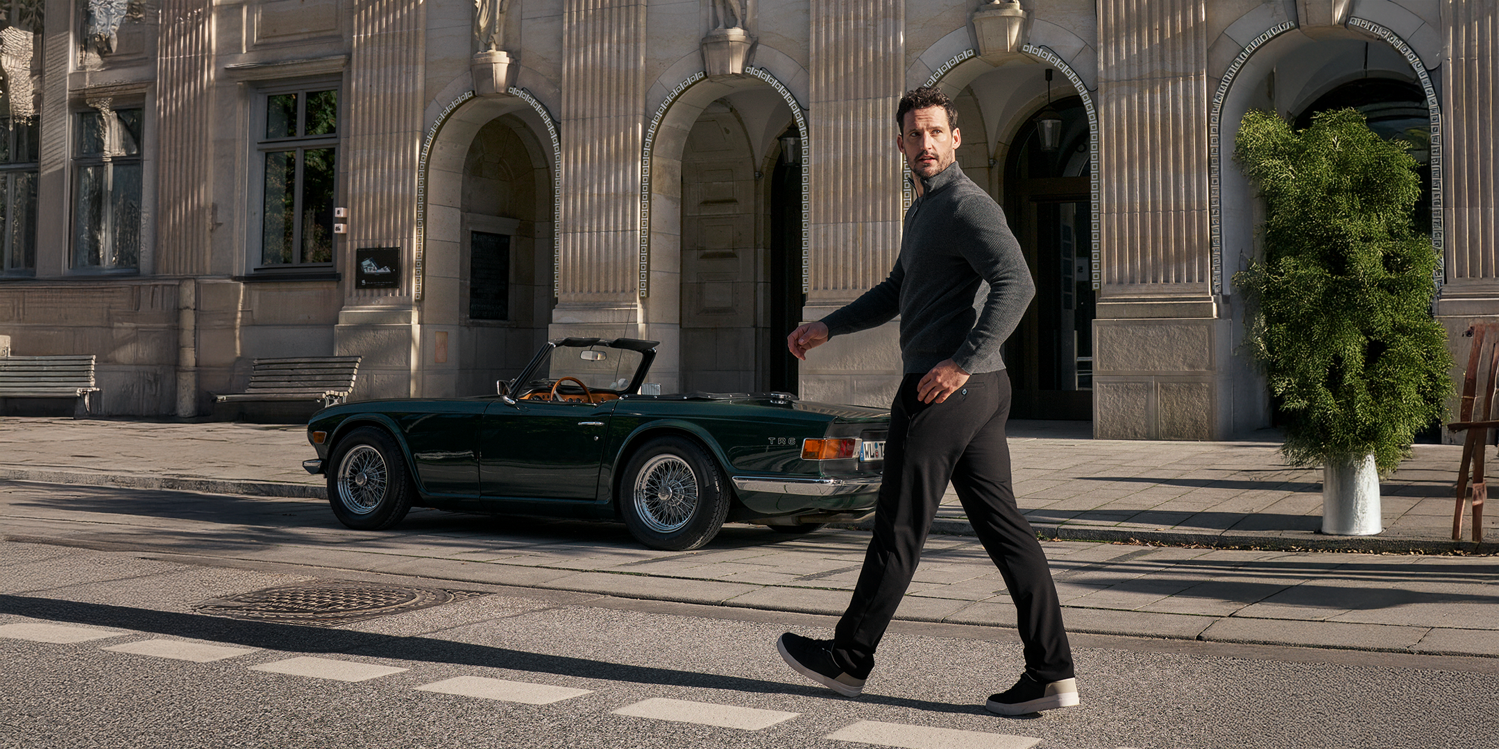 Ein Mann mit dunklem Haar, dunklem Pullover und schwarzen Hosen läuft auf einer Stadtstraße vorbei an einem schwarzen Oldtimer-Cabriolet, das vor einem historischen Gebäude mit großen Fenstern und Steinarchitektur geparkt ist.