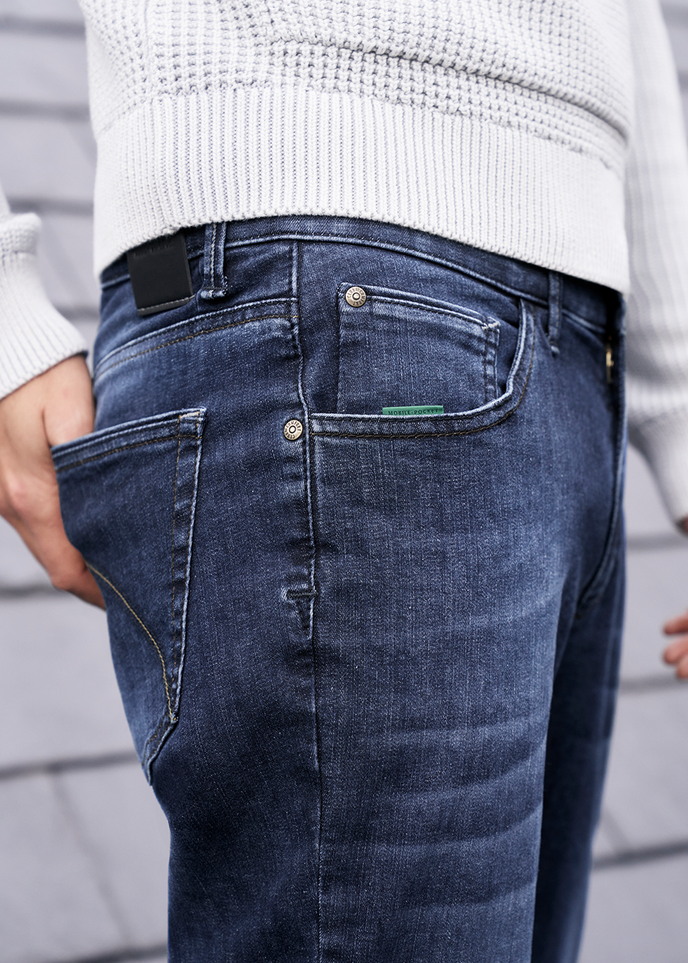 Nahaufnahme eines Mannes, der eine dunkle Jeans trägt. Seine rechte Hand steckt in der Hosentasche. Er trägt außerdem einen hellgrauen Pullover. Der Hintergrund besteht aus einer grauen Holzfassade.