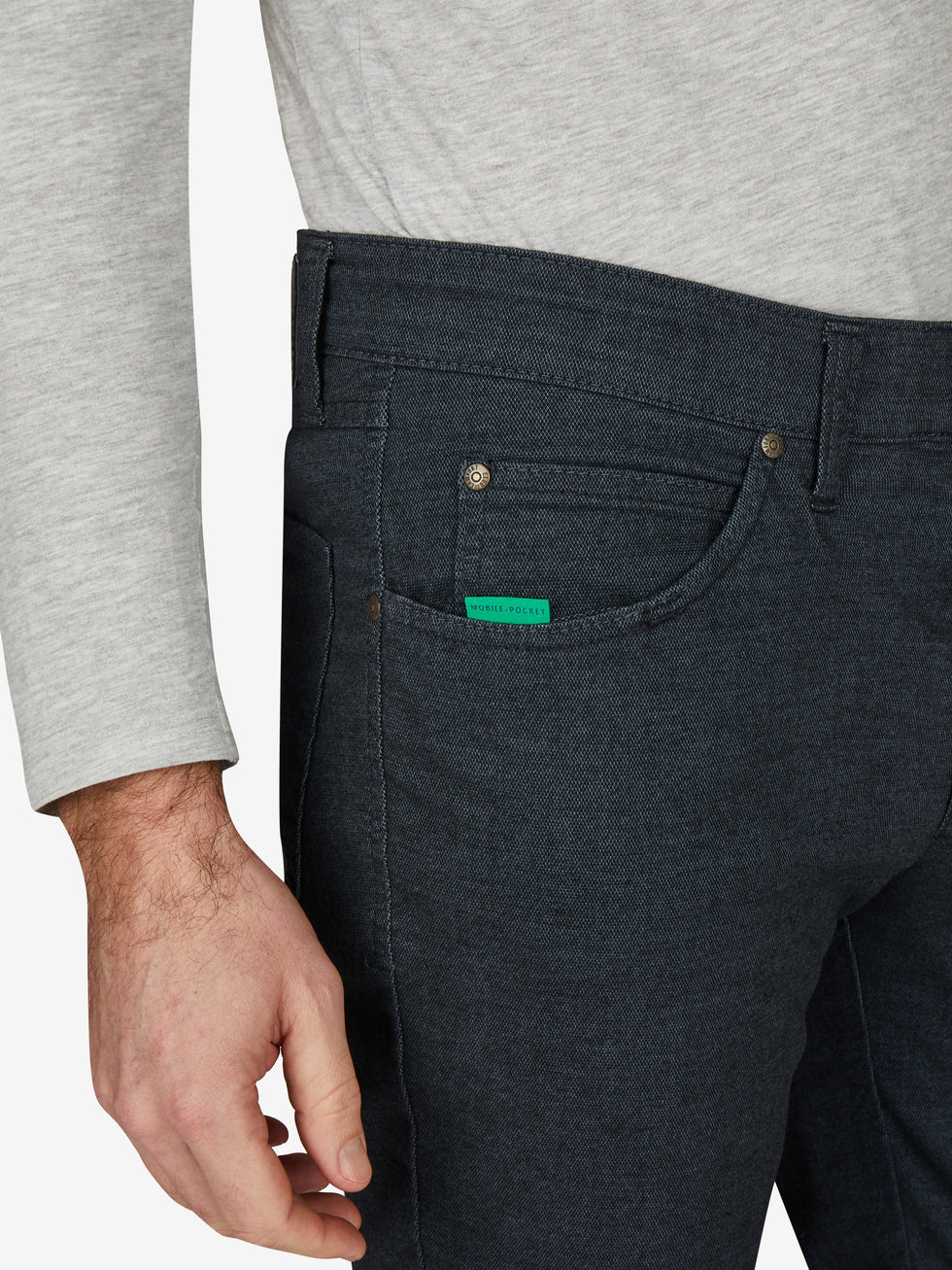 Nahaufnahme einer blauen Baumwollhose "HENRY 7631" in marine melange mit elastischem Doppelstoffbund, Hemdstopper, Münztasche mit Kontrast-Patch, und separater "Mobile Pocket" in der rechten Vordertasche. Dermodel trägt ein graues Oberteil.