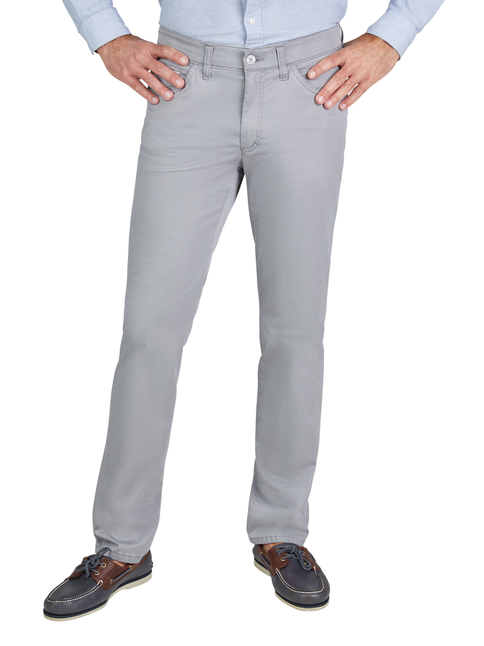 Ein Mann steht mit seinen Händen an den Hüften, trägt eine hellblaue, langärmlige Hemd und helle graue Chino-Hosen. Seine Schuhe sind dunkelblau mit braunen Akzenten und weißer Sohle.