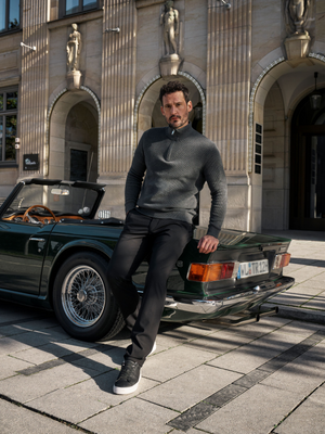 Mann steht vor einem schwarzen Oldtimer, trägt eine dunkelgraue, enganliegende Chino-Hose mit Slim Fit, elastischem Smokbund und Hemdstopper, aus Polyamid-Viskose-Mix in Microstruktur, begleitet von einem dunkelgrauen Pullover, auf einer Stadtstraße.